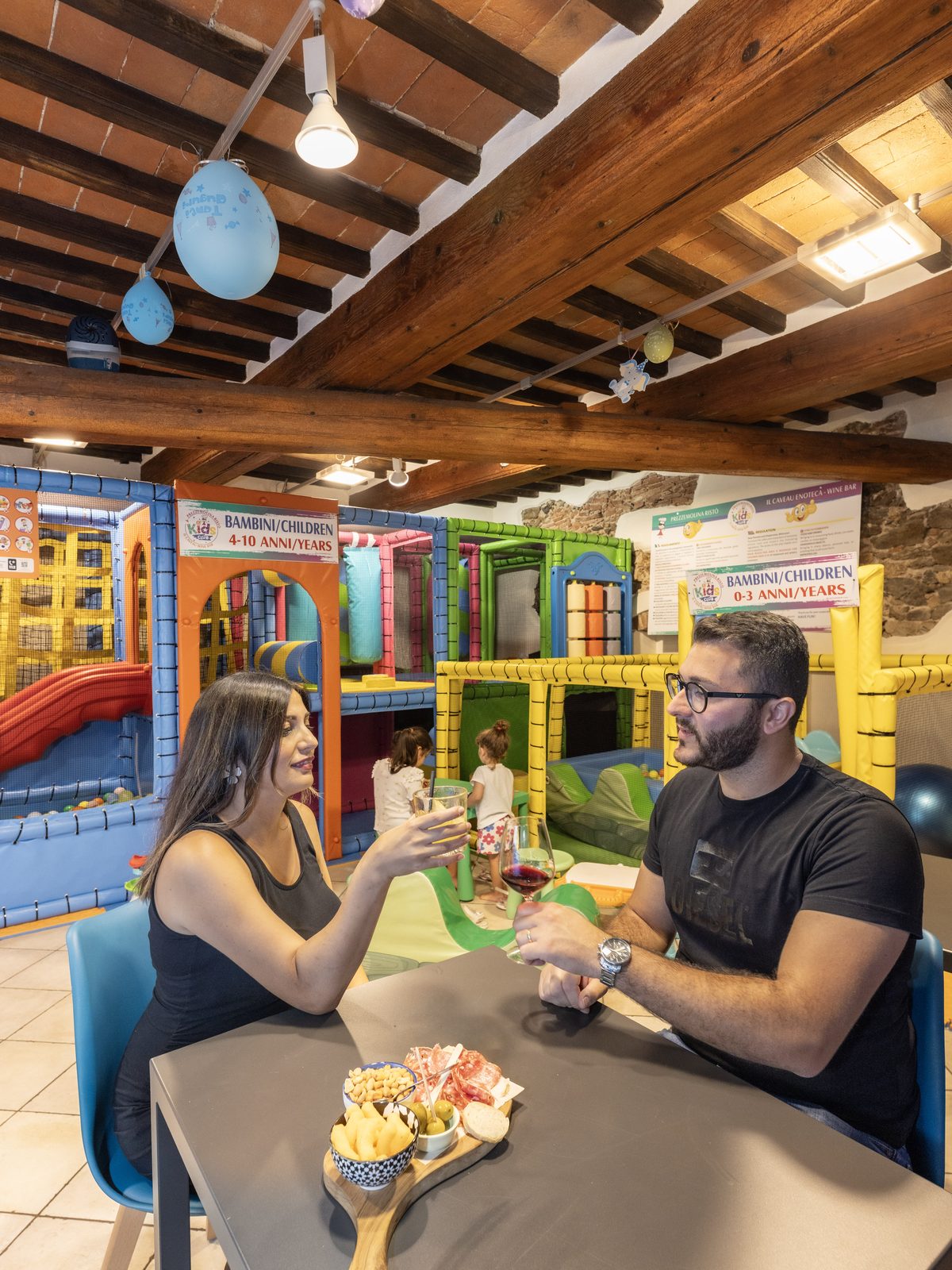 Genitori seduti al tavolo con l'area gioco del Kids Club alle spalle